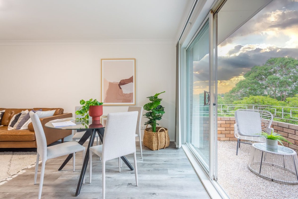 Bright modern apartment dining area with a glass table opening onto a balcony terrace with trees beyond